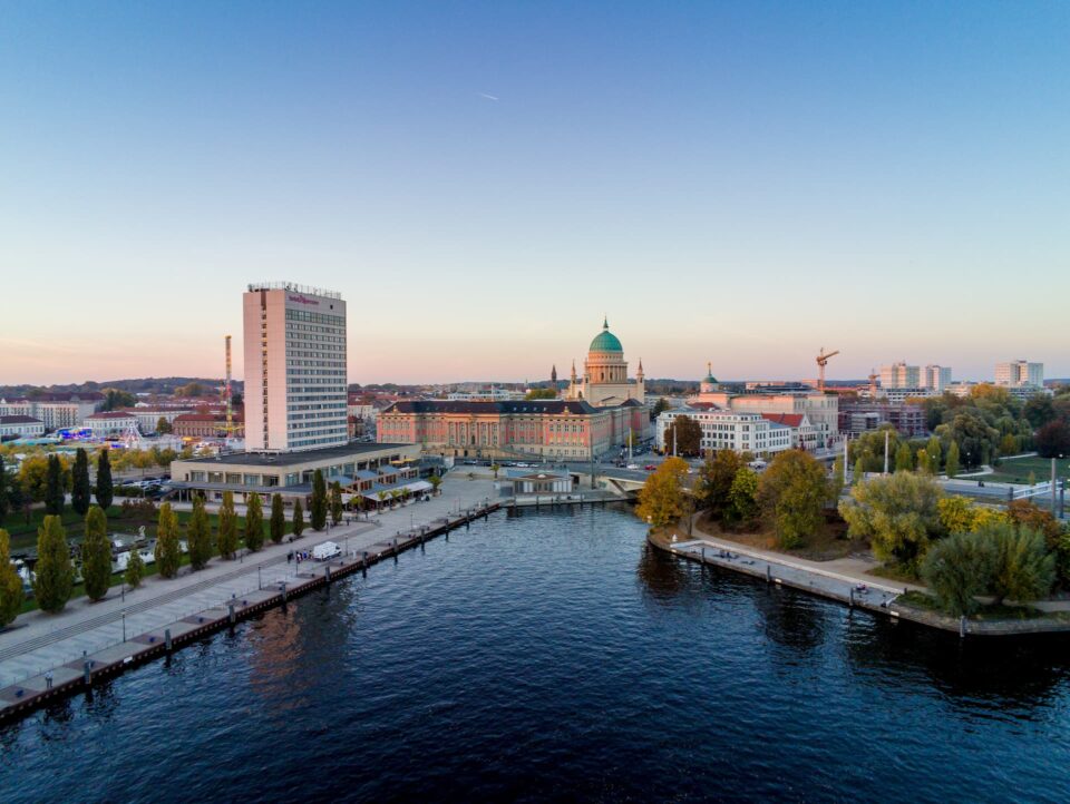 Eine Luftaufnahme der Potsdamer Stadtlandschaft am Wasser bei Sonnenuntergang, die eine Mischung aus moderner und historischer Architektur bietet, die sich perfekt für den Tourismus eignet. Ein historisches Gebäude mit Kuppel sticht zwischen anderen Gebäuden hervor. Bäume mit Herbstlaub säumen die Uferpromenade und eine Promenade erstreckt sich entlang des Wassers.