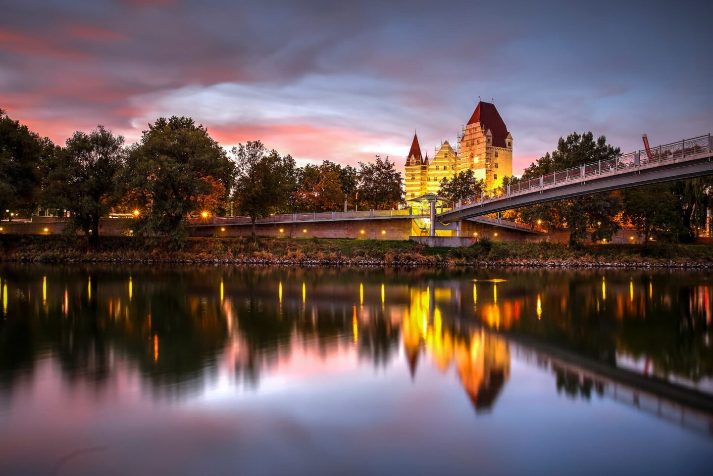 Eine ruhige Abendszene in Ingolstadt zeigt ein beleuchtetes schlossähnliches Gebäude, das sich in einem ruhigen Fluss spiegelt. Eine moderne Brücke erstreckt sich über das Wasser und Bäume säumen das Flussufer unter einem farbenfrohen, wolkenverhangenen Himmel.