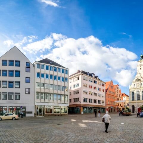 Ein Panoramablick auf einen großen, offenen öffentlichen Platz in Ingolstadt, umgeben von farbenfrohen, historischen Gebäuden in verschiedenen Architekturstilen. Der Himmel ist strahlend blau mit vereinzelten Wolken, und Menschen gehen unter einem sonnigen Himmel spazieren und versammeln sich.