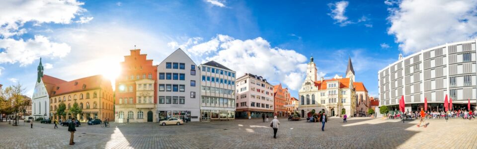 Ein Panoramablick auf einen großen, offenen öffentlichen Platz in Ingolstadt, umgeben von farbenfrohen, historischen Gebäuden in verschiedenen Architekturstilen. Der Himmel ist strahlend blau mit vereinzelten Wolken, und Menschen gehen unter einem sonnigen Himmel spazieren und versammeln sich.