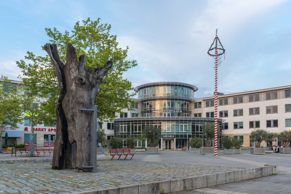 Auf einem Stadtplatz in Ludwigsfelde steht eine große, verwitterte Baumstammskulptur auf einer erhöhten Plattform, die mit Metallstreben befestigt ist. Im Hintergrund ist ein modernes Gebäude mit gläsernem Eingang zu sehen, umgeben von kleineren Gebäuden und Grünflächen. Rechts steht ein Maibaum.