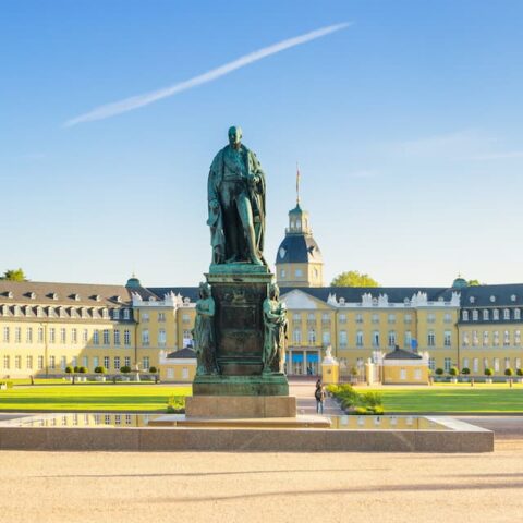 Panoramablick auf das prächtige gelbe Karlsruher Schloss mit einem großen, gepflegten Garten im Vordergrund. In der Mitte ragt eine große Statue einer Person auf einem Sockel hervor. Der Himmel ist klar und blau, mit einer einzelnen weißen Wolke am Himmel. Bäume säumen die Gartenwege.