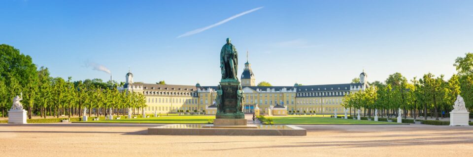 Panoramablick auf das prächtige gelbe Karlsruher Schloss mit einem großen, gepflegten Garten im Vordergrund. In der Mitte ragt eine große Statue einer Person auf einem Sockel hervor. Der Himmel ist klar und blau, mit einer einzelnen weißen Wolke am Himmel. Bäume säumen die Gartenwege.