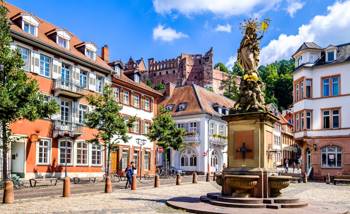 Ein malerischer europäischer Stadtplatz in Heidelberg zeichnet sich durch helle, historische Gebäude mit schrägen Dächern aus. In der Mitte steht ein Brunnen mit einer Statue. Man sieht Bäume, eine Kopfsteinpflasterstraße und Menschen, die einen sonnigen Tag genießen. Im Hintergrund ist ein Schloss auf einem Hügel zu sehen.