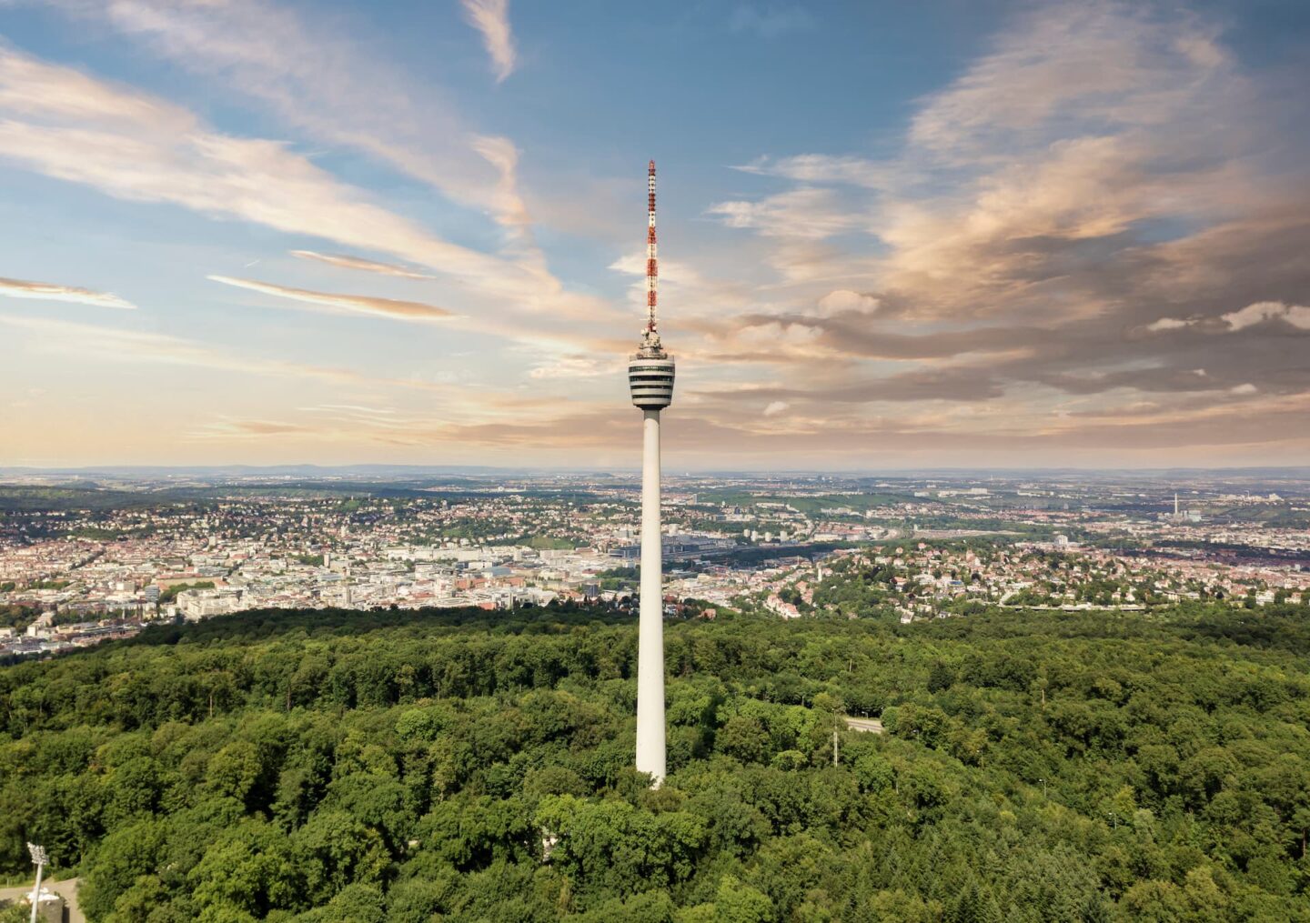 Eine Luftaufnahme eines hohen Fernsehturms mit einer kugelförmigen Aussichtsplattform erhebt sich über dem üppigen grünen Wald in der Nähe von Stuttgart. Im Hintergrund erstreckt sich die Stadtlandschaft unter einem teilweise bewölkten Himmel und zeigt eine gewaltige Stadtentwicklung.