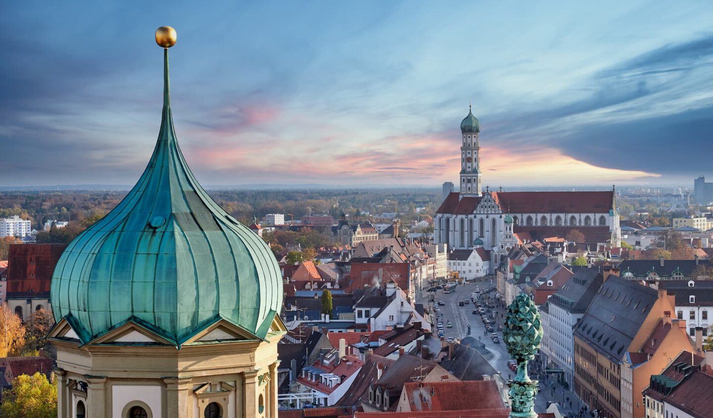 Das Foto zeigt einen Panoramablick auf Augsburg, Deutschland, mit der verzierten grünen Kuppel der St. Ulrichskirche im Vordergrund. In der Ferne erhebt sich der hohe Turm des Augsburger Rathauses vor dem Hintergrund eines farbenfrohen Sonnenuntergangshimmels und hebt die historische Skyline von Augsburg hervor.