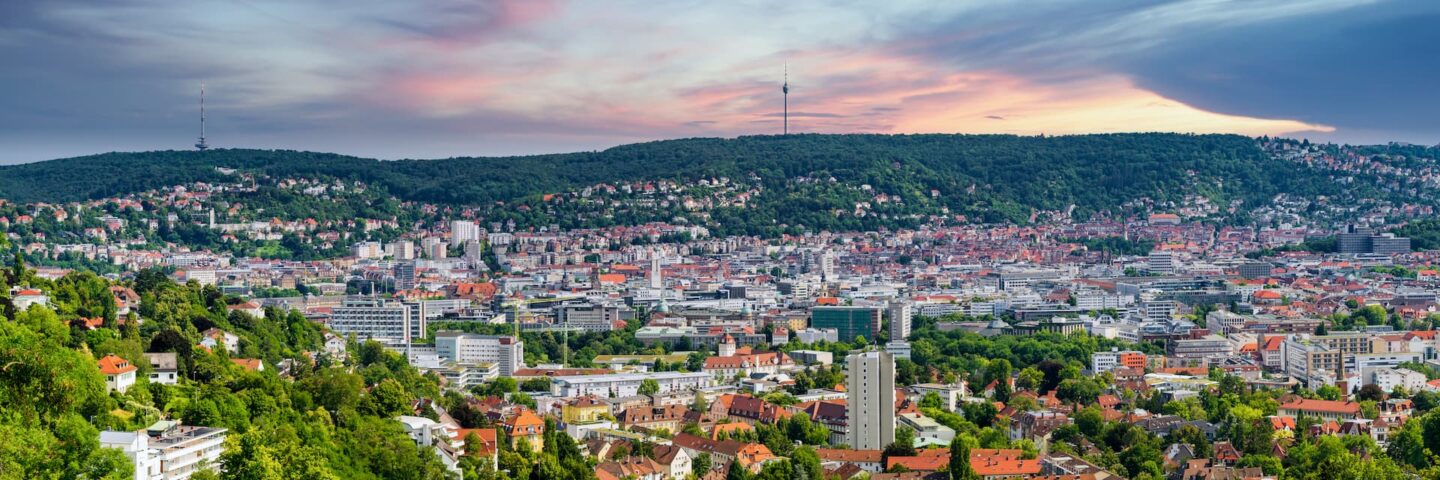 Panoramablick auf Stuttgart bei Sonnenuntergang mit einer Mischung aus modernen und traditionellen Gebäuden, umgeben von üppigen grünen Hügeln. Die Skyline zeichnet sich durch einige hohe Gebäude aus und der Himmel ist in Blau-, Lila- und Rosatönen gefärbt.