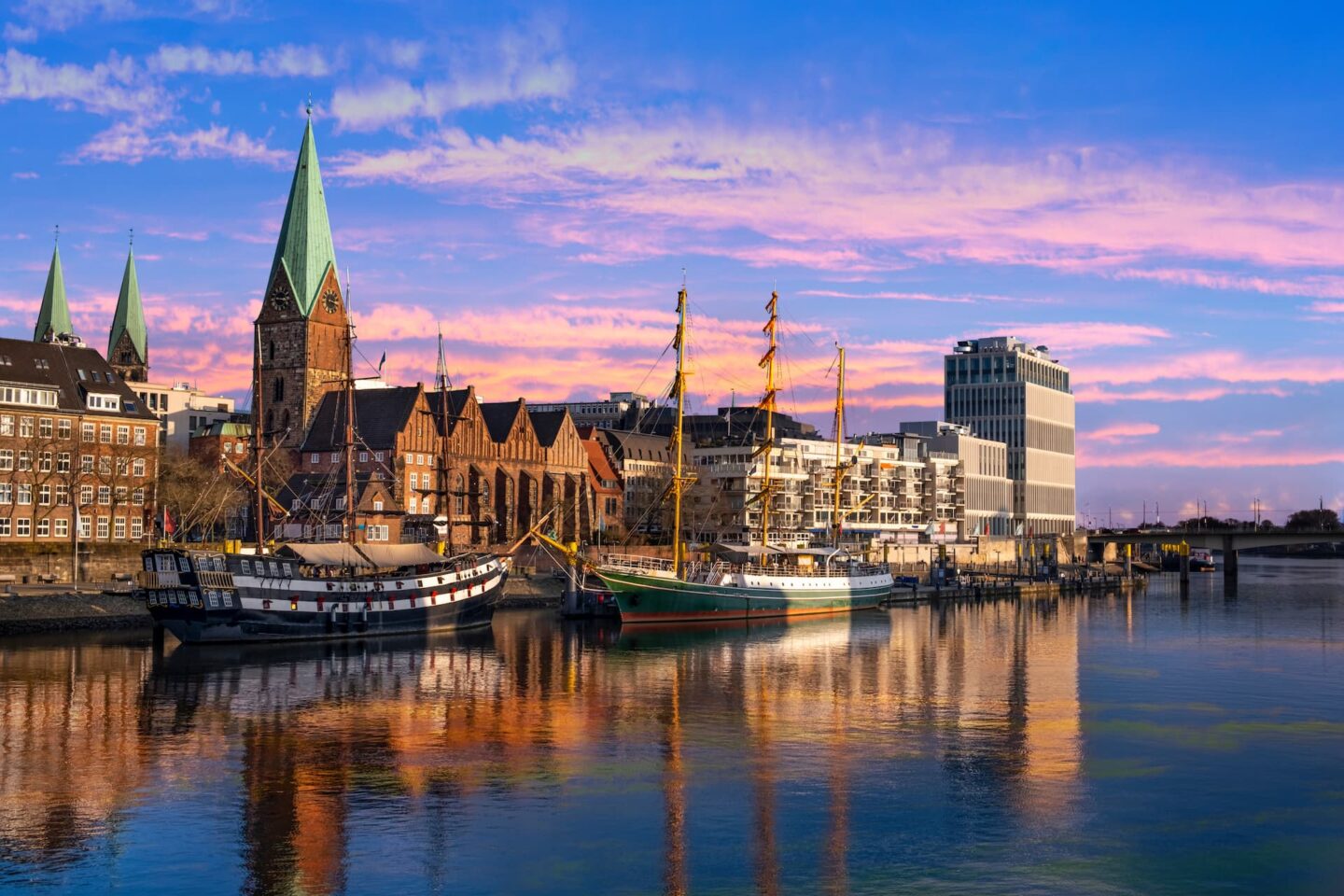 Eine malerische Hafenszene bei Sonnenuntergang in Bremen zeigt große Schiffe, die neben historischen Gebäuden mit Backsteinfassaden und grünen Kirchtürmen anlegen und sich im ruhigen Wasser spiegeln. Moderne Gebäude stehen im Kontrast zur historischen Architektur unter einem lebendigen, farbenfrohen Himmel.