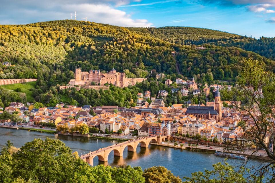 Eine malerische Aussicht auf Heidelberg, Deutschland, mit seinen ikonischen Gebäuden mit roten Dächern, dem stattlichen Heidelberger Schloss auf einem Hügel und der bezaubernden Alten Brücke über den Neckar. Umgeben von üppigem Grün und unter einem strahlend blauen Himmel fängt diese Szene Heidelbergs zeitlose Schönheit ein.