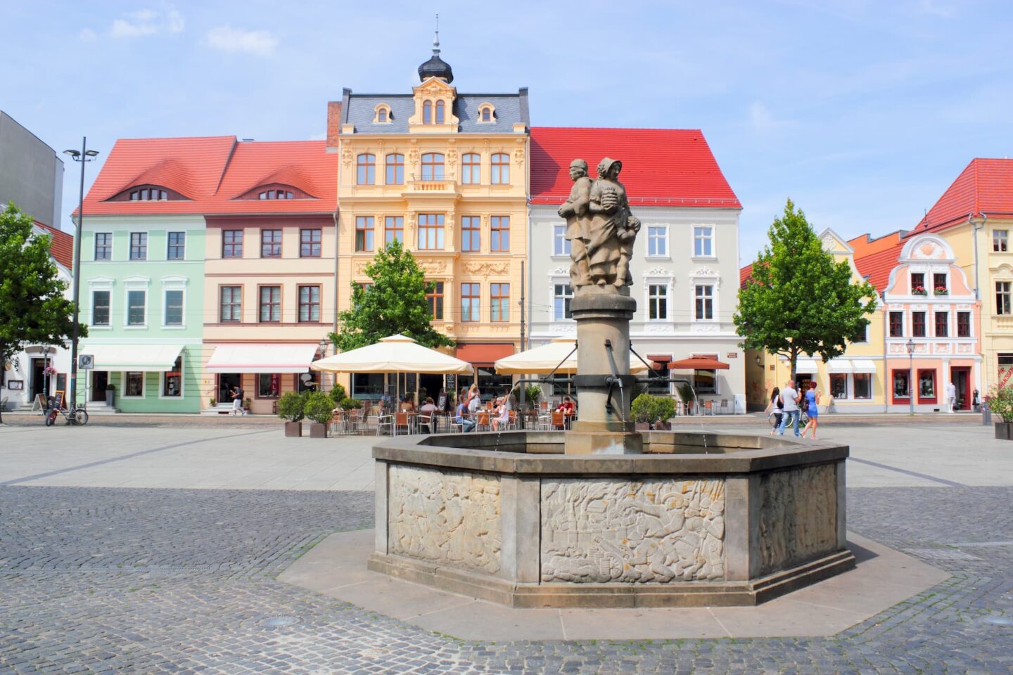 Auf einem malerischen Stadtplatz in Cottbus steht in der Mitte ein Steinbrunnen mit einer Statue. Der Platz ist von farbenfrohen historischen Gebäuden mit roten und orangefarbenen Dächern umgeben. Sitzgelegenheiten im Freien von Cafés säumen den Platz, und ein paar Leute schlendern gemütlich umher und sitzen herum.