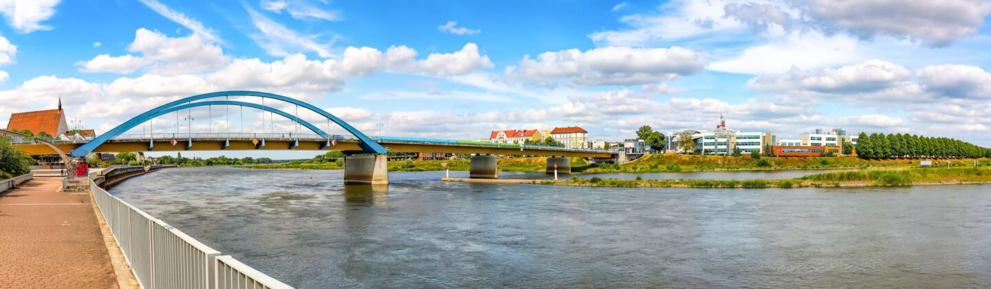 Panoramablick auf eine große blaue Brücke, die sich über die breite Oder spannt, unter einem klaren, blauen Himmel mit vereinzelten Wolken. Auf der einen Seite gibt es einen Fußgängerweg, während auf der anderen Seite Gebäude und Bäume das Frankfurter Flussufer säumen. Dieser malerische Ort ist perfekt für den Tourismus und bietet eine ruhige und helle Atmosphäre.