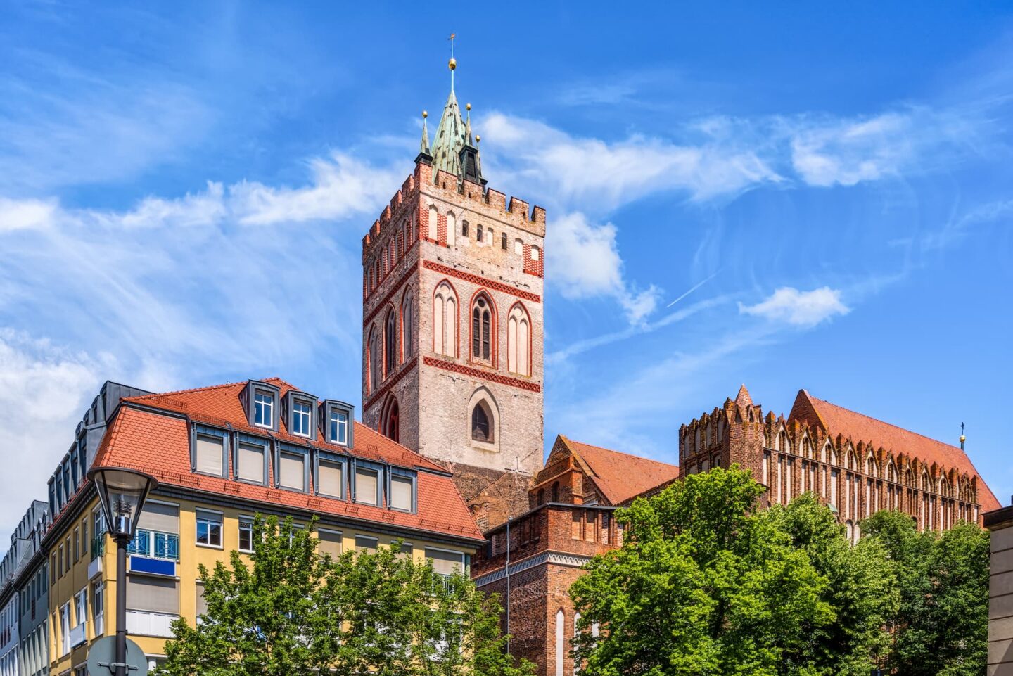 Ein hoher, historischer Backsteinturm im gotischen Stil erhebt sich vor einem strahlend blauen Himmel in Frankfurt, Deutschland. Flankiert von kleineren Gebäuden mit Ziegeln, Schindeln und Grünanlagen zeichnet sich die mittelalterliche Architektur durch komplizierte Details, gewölbte Fenster und einen spitzen Turm mit einer Wetterfahne an der Spitze aus.