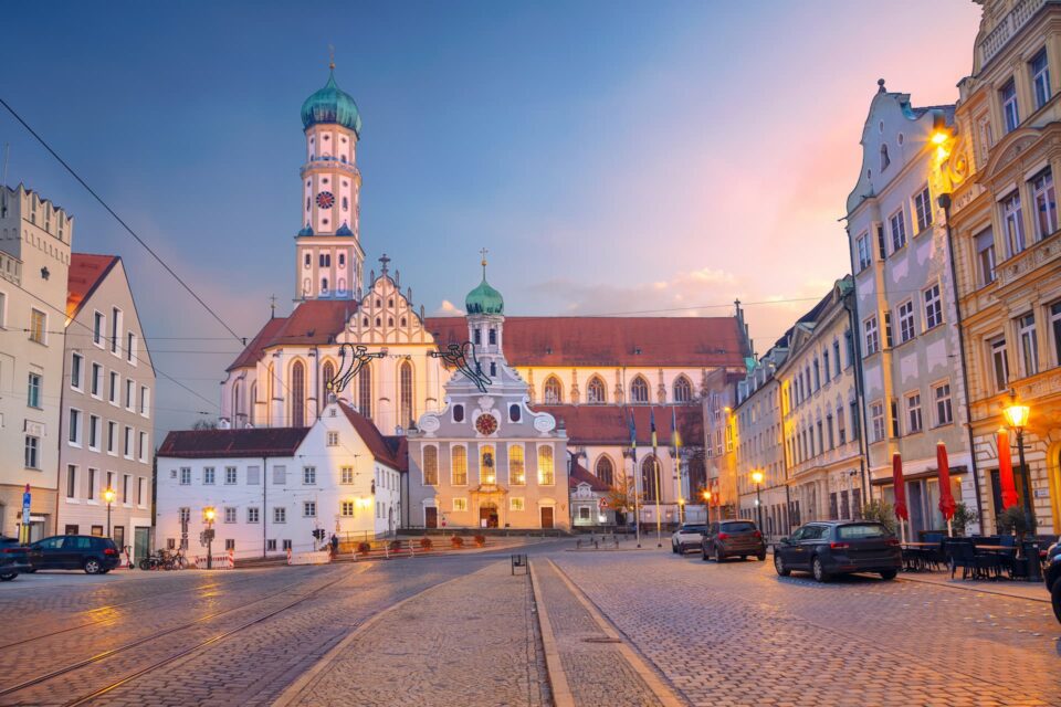 Eine malerische Ansicht von Augsburg, Deutschland, mit Kopfsteinpflasterstraßen und historischer Architektur. Das Bild zeigt die Abtei St. Ulrich und Afra bei Sonnenuntergang mit ihrer kunstvollen Fassade und den Zwillingstürmen. Die umliegenden Augsburger Gebäude und Straßenlaternen tragen zur ruhigen Atmosphäre bei.