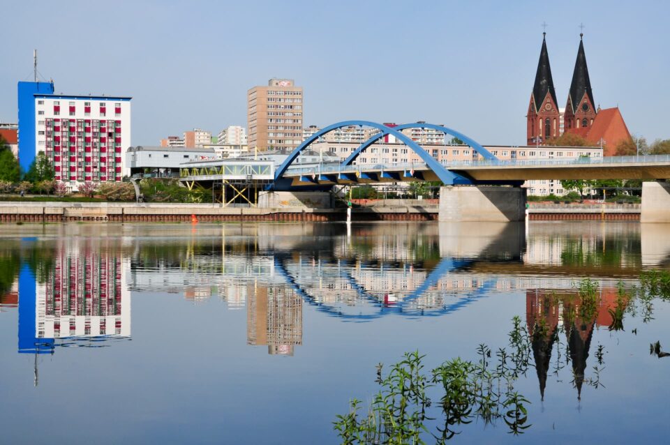 Eine Stadtlandschaft mit einer blau gewölbten Brücke über der ruhigen Oder, in der sich Brücke und Gebäude spiegeln. Im Hintergrund sieht man eine Mischung aus modernen Wohnungen und einer Kirche mit zwei Türmen in Frankfurt, Deutschland. Das Flussufer ist von Grünflächen gesäumt, die einen natürlichen Kontrast zur städtischen Umgebung bilden.