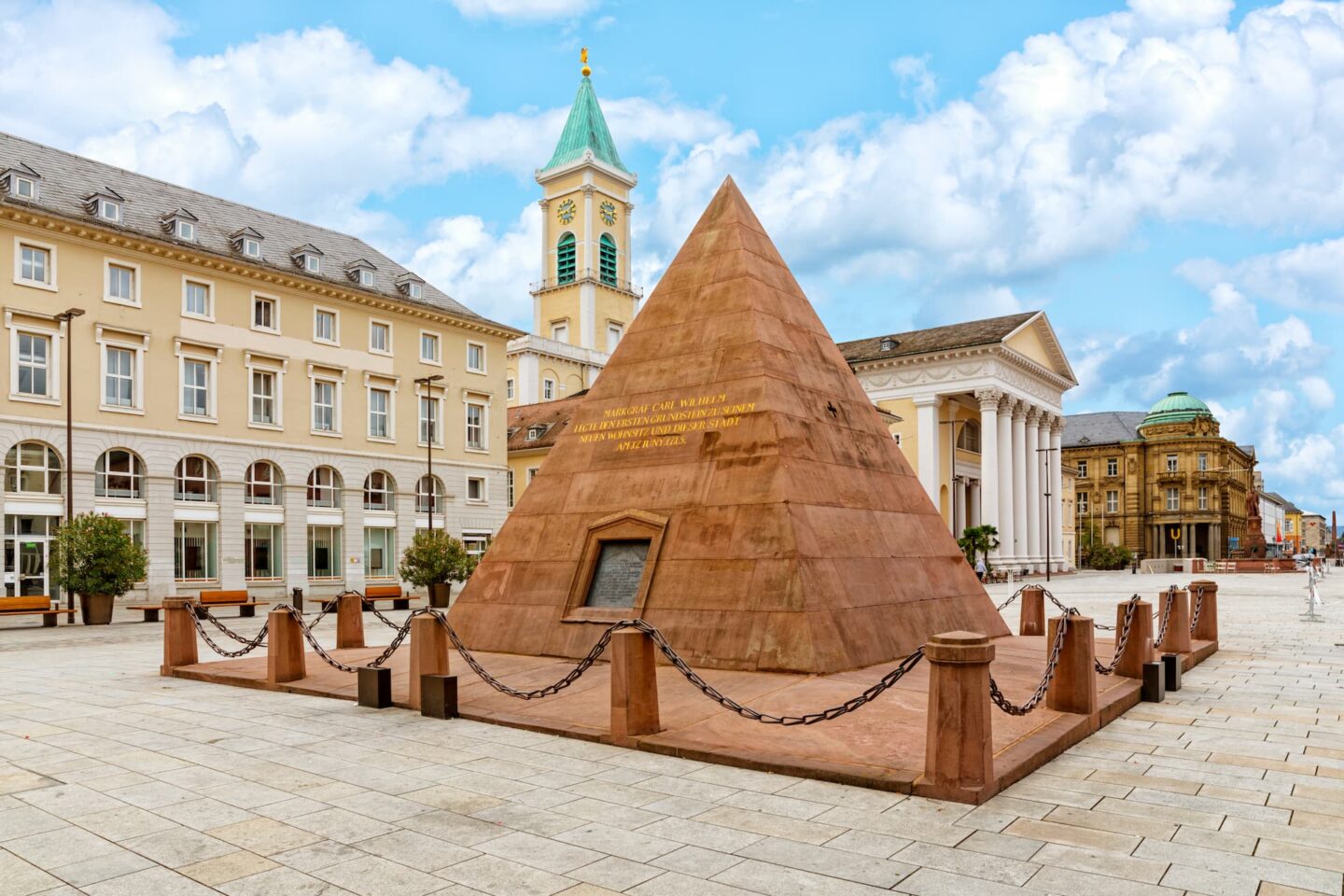 In der Mitte eines gepflasterten Platzes in Karlsruhe steht ein Pyramidendenkmal aus rotem Sandstein, umgeben von Ketten. In der Nähe stehen beige-weiße Gebäude, darunter eines mit einem Turm mit grüner Kuppel. Der Himmel darüber ist teilweise bewölkt.