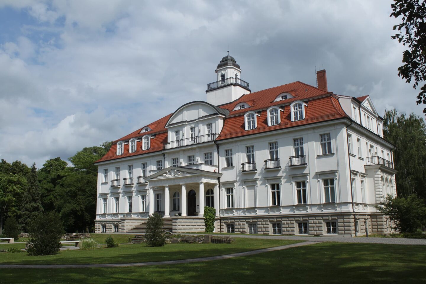 An einem sonnigen Tag sieht man ein großes, elegantes weißes Herrenhaus in Ludwigsfelde mit einem roten Ziegeldach und zahlreichen Fenstern. Das Gebäude verfügt über einen hohen zentralen Turm und einen reich verzierten Eingang mit Säulen. Es ist von einem üppigen, gepflegten Rasen und Bäumen umgeben, über dem sich ein teilweise bewölkter Himmel befindet.