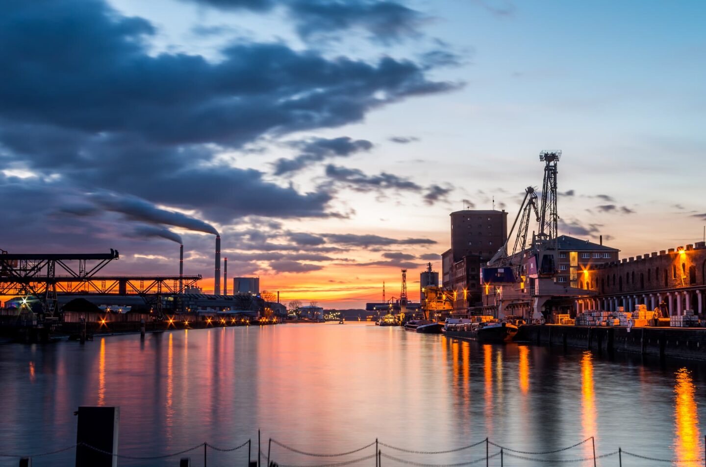 Ein malerischer Blick auf den Karlsruher Industriehafen bei Sonnenuntergang. Der Himmel ist in leuchtenden Orange-, Rosa- und Blautönen gehalten. Industrielle Strukturen, darunter ein Dock, Kräne und ein rauchender Schornstein, säumen den Wasserrand, während das Wasser den farbenfrohen Himmel widerspiegelt.