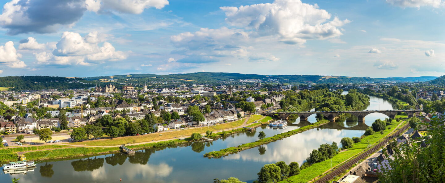 Ein Panoramablick auf Trier, eingebettet in einen gewundenen Fluss. Die Szene ist üppig grün, mit blauem Himmel und vereinzelten Wolken. Brücken überspannen den Fluss und verbinden verschiedene Teile der Stadt, während Hügel und historische Gebäude den Hintergrund füllen.