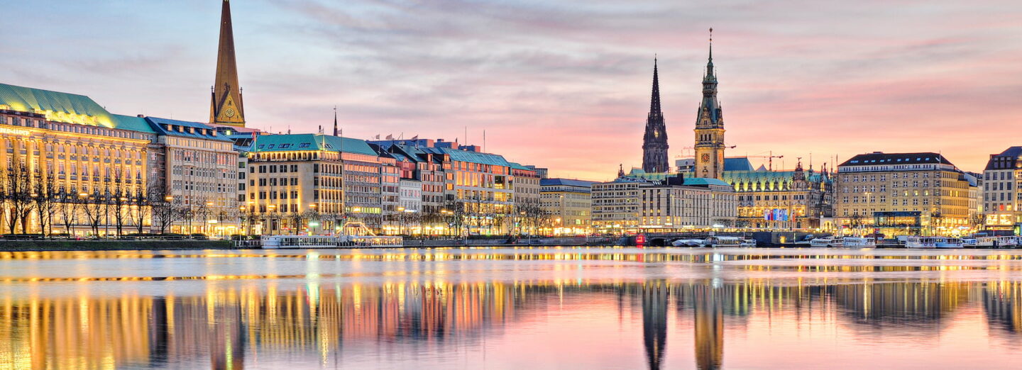 Ein Panoramablick auf Hamburg bei Sonnenuntergang zeigt den Alstersee mit klaren Spiegelungen historischer Gebäude und Kirchtürme. Der Himmel ist in Rosa- und Orangetönen gefärbt und unterstreicht das malerische Stadtbild.