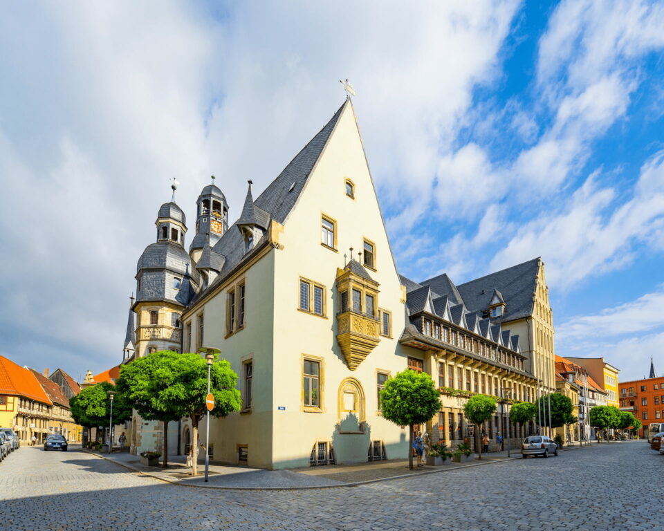 Ein malerischer Blick auf den historischen Stadtplatz von Aschersleben zeigt ein markantes gelbes Gebäude mit einem steilen Satteldach und kunstvollen architektonischen Details. Weitere charmante Gebäude säumen die gepflasterte Straße, alle unter einem hellen Himmel mit vereinzelten Wolken. Üppige grüne Bäume sorgen für lebendige Farbe.