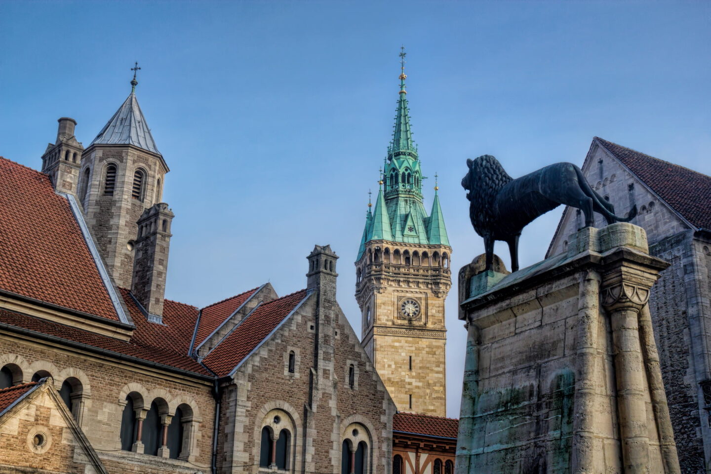 Eine Architekturszene mit historischen Gebäuden mit Steinfassaden und roten Ziegeldächern, die an Braunschweig erinnern. Im Hintergrund ragt ein zentraler Turm mit einer grünen Turmspitze und einer Uhr empor, während im Vordergrund eine bronzene Löwenstatue auf einem Steinsockel thront. Der Himmel ist klar und blau.