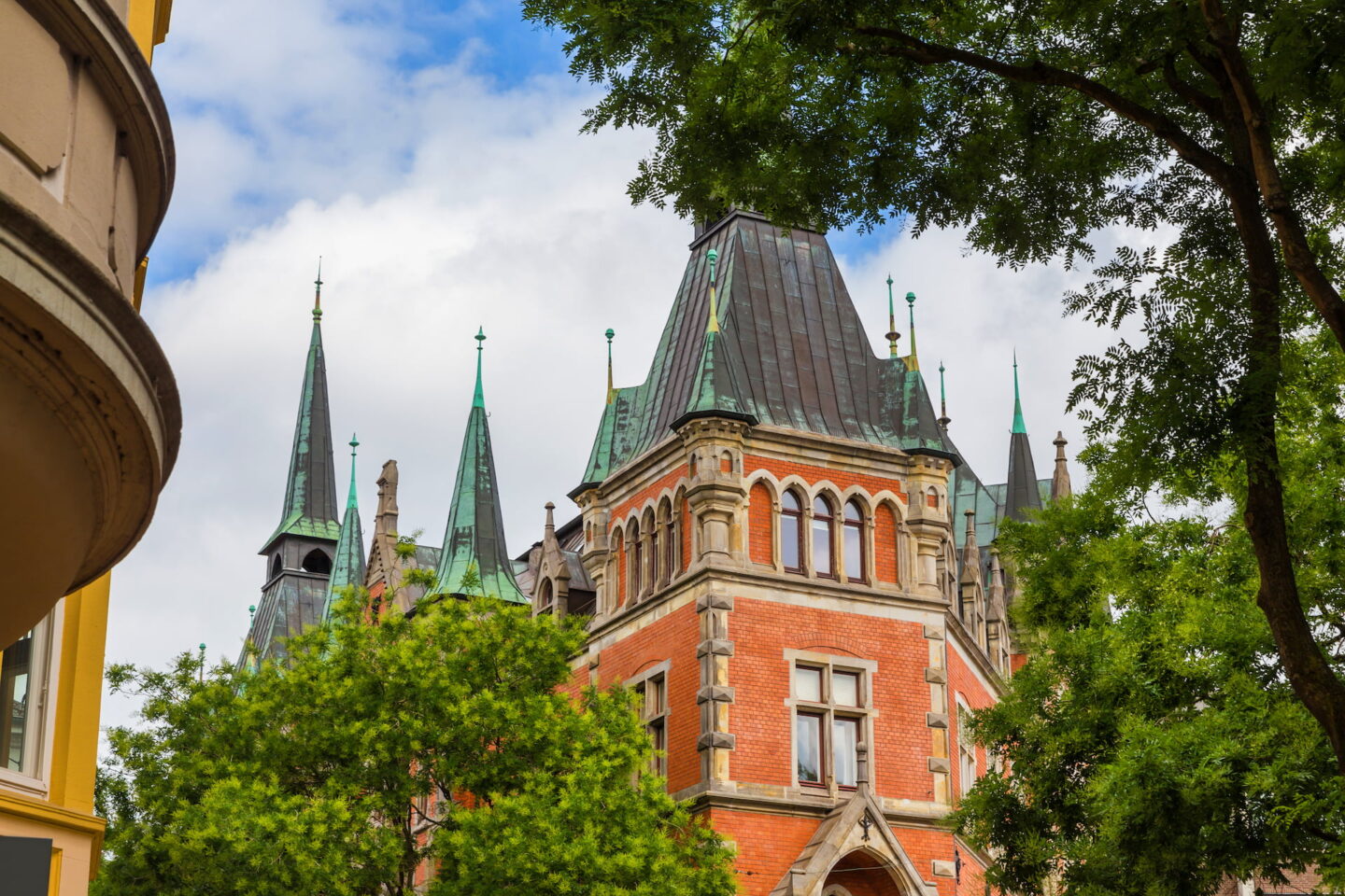 Ein historisches Gebäude in Oldenburg mit roten Backsteinmauern, Steinakzenten und einem hohen, dunklen Dach mit mehreren Türmen wird von grünen Bäumen eingerahmt. Der Himmel ist teilweise bewölkt und auf der linken Seite des Bildes ist eine moderne Gebäudeecke zu sehen.