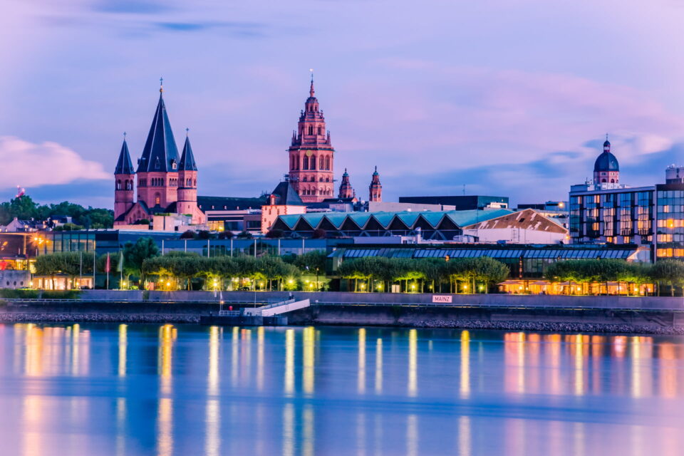 Ein ruhiger Blick auf Mainz in der Abenddämmerung zeigt seine historische Architektur. Hohe Kirchtürme und eine markante Kathedrale heben sich von einem schwach beleuchteten Himmel ab. Der ruhige Fluss im Vordergrund spiegelt die beleuchteten Gebäude und trägt zur malerischen Szene bei.