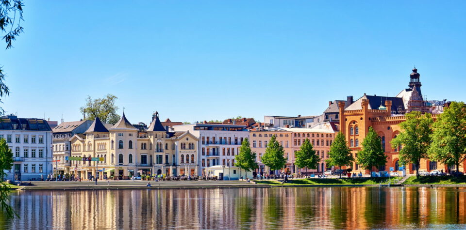 Ein malerischer Blick auf Schwerin am Seeufer mit charmanten, historischen Gebäuden in unterschiedlichen Architekturstilen. Bäume säumen das Ufer und der klare blaue Himmel spiegelt sich im ruhigen Wasser. Man sieht Menschen, die entlang der Schweriner Seepromenade spazieren.