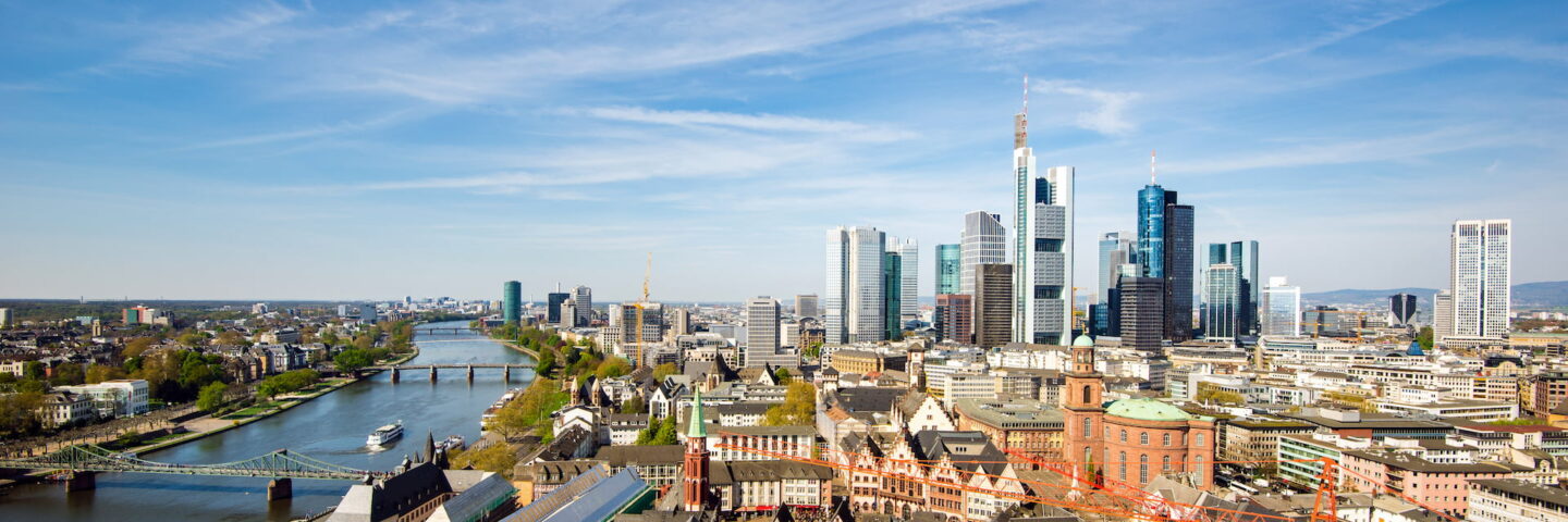 Ein weitläufiger Blick auf Frankfurt am Main offenbart eine Stadtlandschaft, durch die ein Fluss fließt. Die Gegend besticht durch eine Mischung aus historischen Gebäuden und modernen Wolkenkratzern, mehreren Brücken über den Fluss und einem klaren blauen Himmel, der von üppigem Grün umrahmt wird.