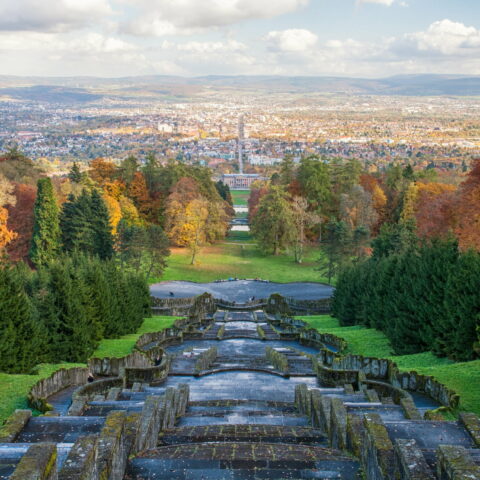 Ein Panoramablick von einem hohen Aussichtspunkt in Kassel überblickt die Stadt in der Ferne, mit einer absteigenden Treppe im Vordergrund. Die Szene wird von Bäumen eingerahmt, die Herbstfarben zeigen – Orange-, Rot- und Grüntöne – unter einem klaren Himmel mit vereinzelten Wolken darüber.