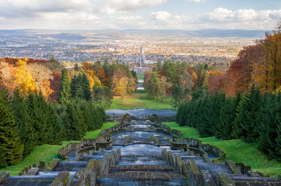 Ein Panoramablick von einem hohen Aussichtspunkt in Kassel überblickt die Stadt in der Ferne, mit einer absteigenden Treppe im Vordergrund. Die Szene wird von Bäumen eingerahmt, die Herbstfarben zeigen – Orange-, Rot- und Grüntöne – unter einem klaren Himmel mit vereinzelten Wolken darüber.