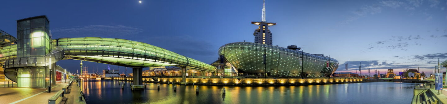 Panoramablick auf eine moderne Hafenstadtlandschaft in Bremerhaven bei Abenddämmerung mit einem beleuchteten futuristischen Gebäude und einer gläsernen Fußgängerbrücke über dem Wasser. Der Himmel wechselt von tiefem Blau zu hellem Blau mit sanften Spiegelungen auf der ruhigen Wasseroberfläche.