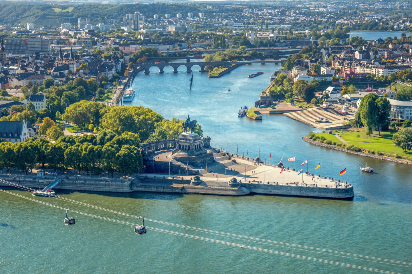 Luftaufnahme des Deutschen Ecks in Koblenz, Deutschland, wo die Mosel auf den Rhein trifft. Das monumentale Reiterstandbild von Kaiser Wilhelm I. ist zu sehen. Die Umgebung ist üppig mit Bäumen, Gebäuden und zwei parallelen Seilbahnen, die den Fluss in Koblenz überqueren.