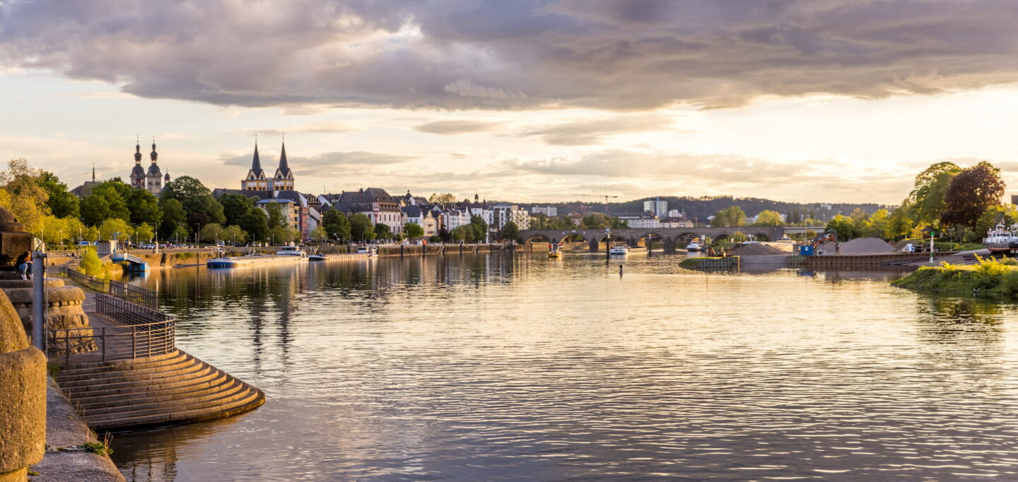 Malerische Aussicht auf die europäische Flussstadt Koblenz bei Sonnenuntergang mit historischen Gebäuden mit Türmen, einer Steinbrücke über den Fluss, am Flussufer angedockten Booten und üppigem Grün am Wasser. Der Himmel ist von dramatischen Wolken mit warmem, goldenem Licht geprägt.