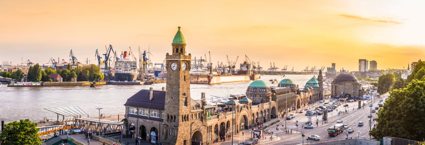 Panoramablick auf die Landungsbrücken in Hamburg bei Sonnenuntergang, mit dem Glockenturm und dem geschäftigen Hafen im Hintergrund. Das Bild fängt die dynamische Mischung aus historischer Architektur, belebten Straßen und Industriekränen entlang dieser lebhaften Hafenpromenade ein.