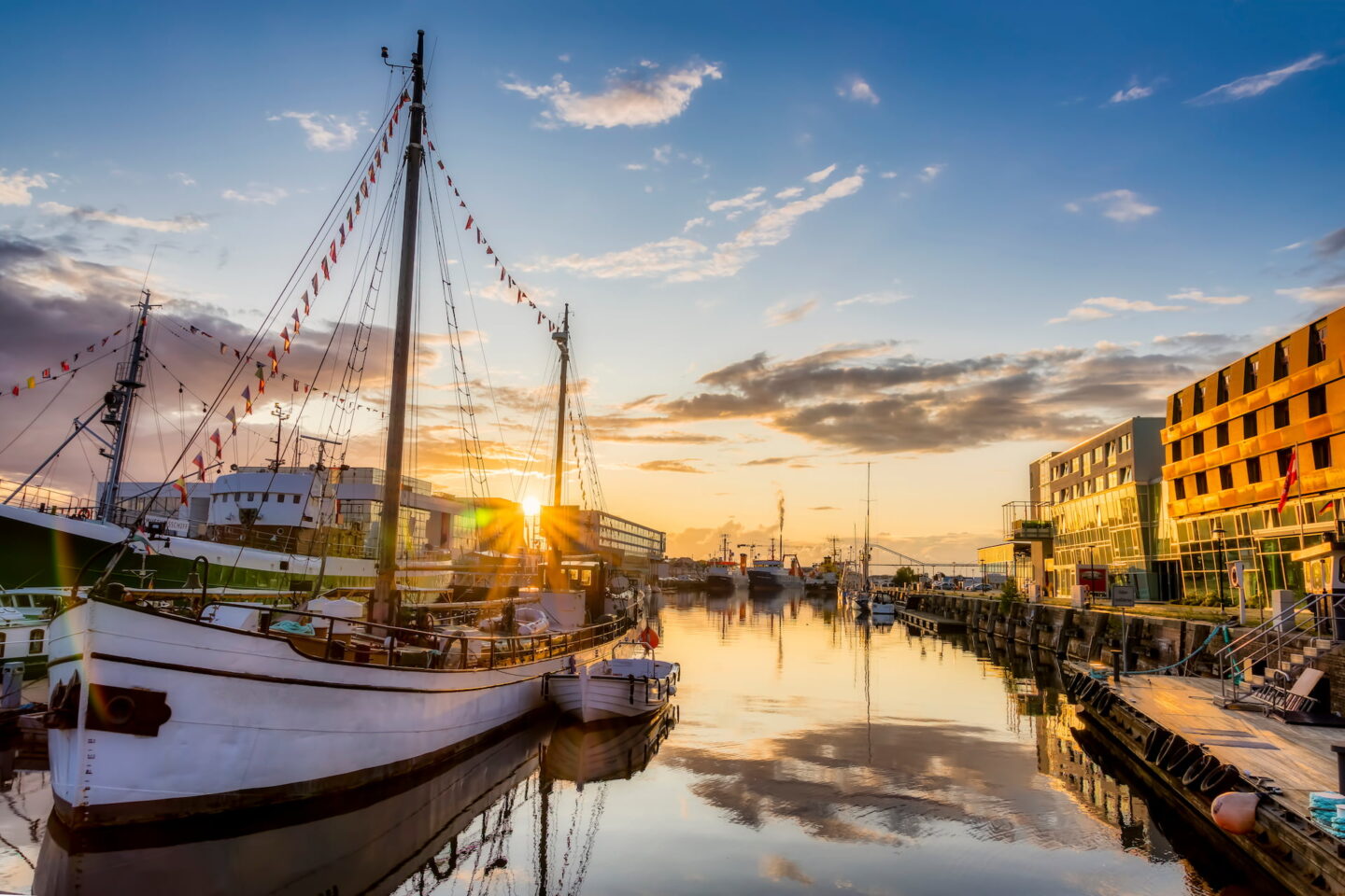 Eine ruhige Hafenszene bei Sonnenuntergang mit Booten, die am ruhigen Wasser angedockt sind. Der Himmel ist mit Wolken geschmückt, die das goldene Licht der untergehenden Sonne reflektieren. Moderne Gebäude säumen die rechte Seite des Bremerhavener Hafens, während auf der gegenüberliegenden Seite einige größere Schiffe zu sehen sind.