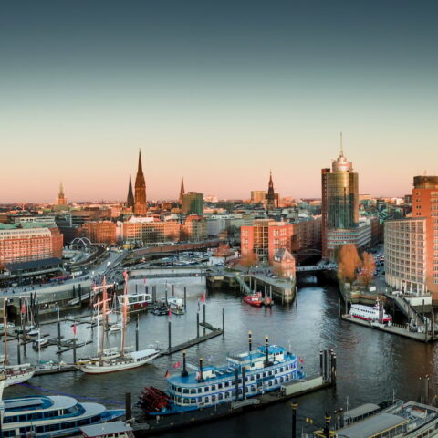 Ein Panoramablick auf die Hamburger Hafenstadt bei Sonnenuntergang zeigt am Wasser angedockte Boote, moderne und historische Gebäude sowie einen prominenten Konzertsaal mit einer einzigartigen Glasfassade, die das Abendlicht reflektiert. Der Himmel ist klar und weist einen Farbverlauf von hellen zu dunkleren Farbtönen auf.