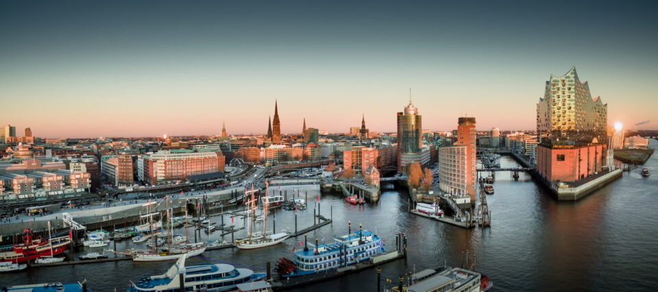 Ein Panoramablick auf die Hamburger Hafenstadt bei Sonnenuntergang zeigt am Wasser angedockte Boote, moderne und historische Gebäude sowie einen prominenten Konzertsaal mit einer einzigartigen Glasfassade, die das Abendlicht reflektiert. Der Himmel ist klar und weist einen Farbverlauf von hellen zu dunkleren Farbtönen auf.