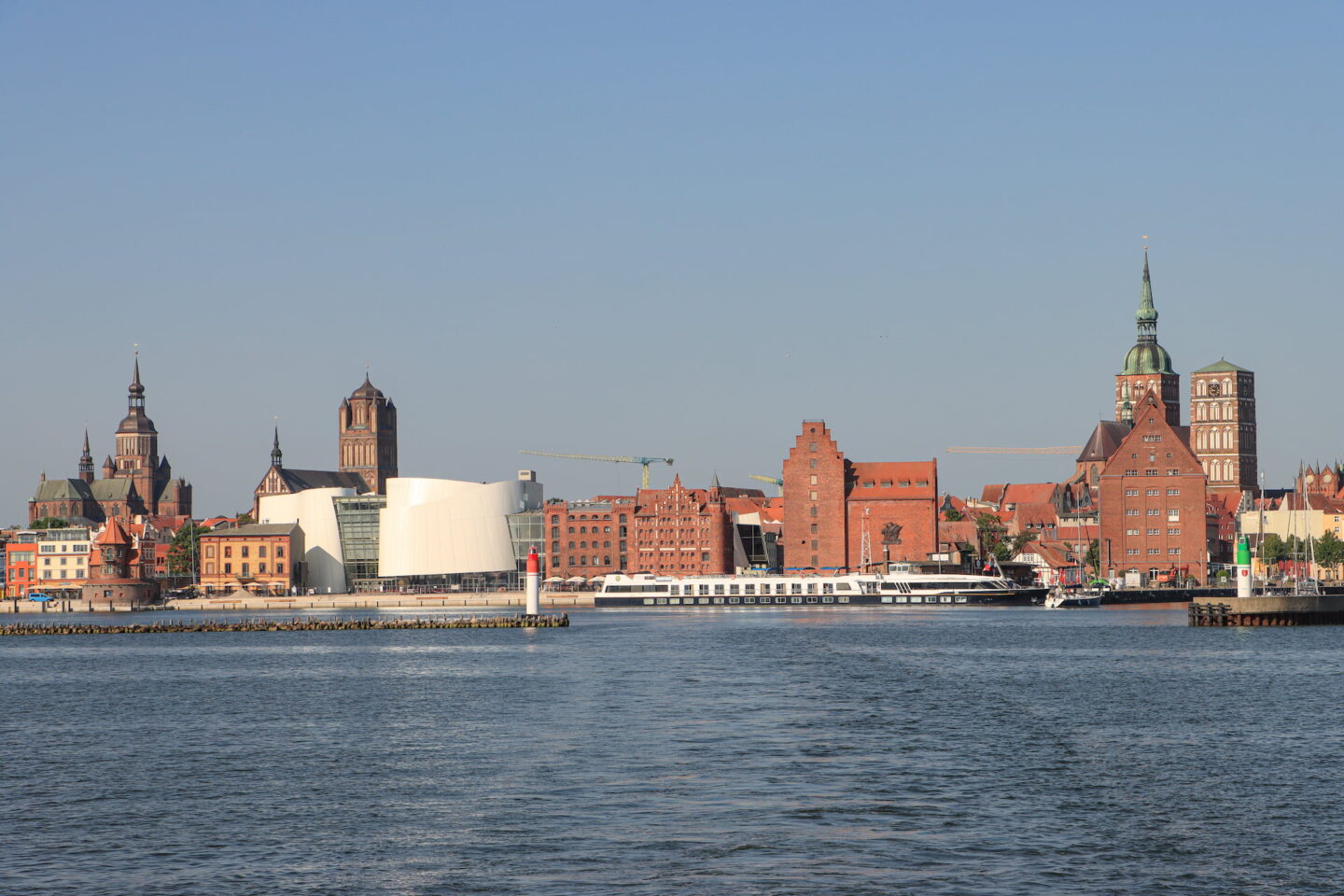 Ein malerischer Blick auf die Uferpromenade der historischen Stadt Stralsund mit markanten Backsteingebäuden und roten Dächern, Kirchtürmen und einem weißen modernen Gebäude. Ein Boot liegt am Ufer und im Vordergrund erstreckt sich das ruhige Wasser unter einem klaren blauen Himmel.