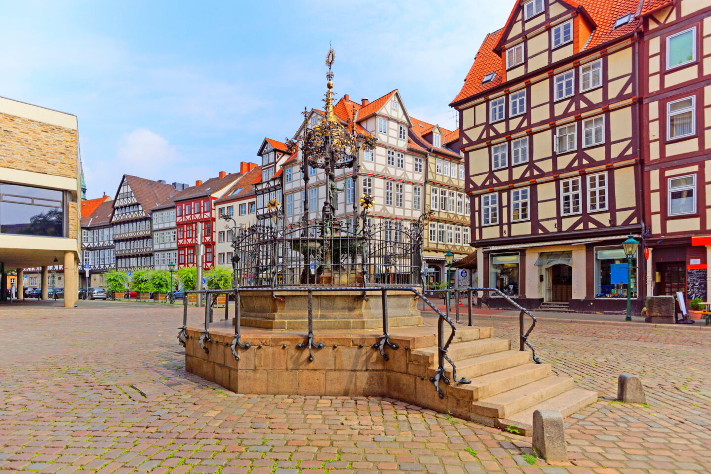 Ein historischer Platz in Hannover mit Kopfsteinpflaster verfügt über einen dekorativen Brunnen mit schmiedeeisernen Geländern und Stufen. Um den Brunnen herum stehen Fachwerkhäuser mit roten Dächern und weißen Fenstern, die die klassische Architektur der Gegend zur Schau stellen.