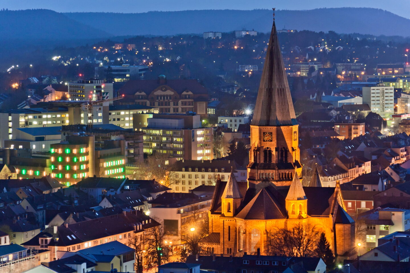 Eine malerische Aussicht auf das historische Kaiserslautern in der Abenddämmerung, mit einer markanten beleuchteten Kirche mit einem hohen Turm im Vordergrund. Das Stadtbild vereint moderne Gebäude und Häuser, die sich vor Hügeln unter einem dunkelblauen Himmel abheben.