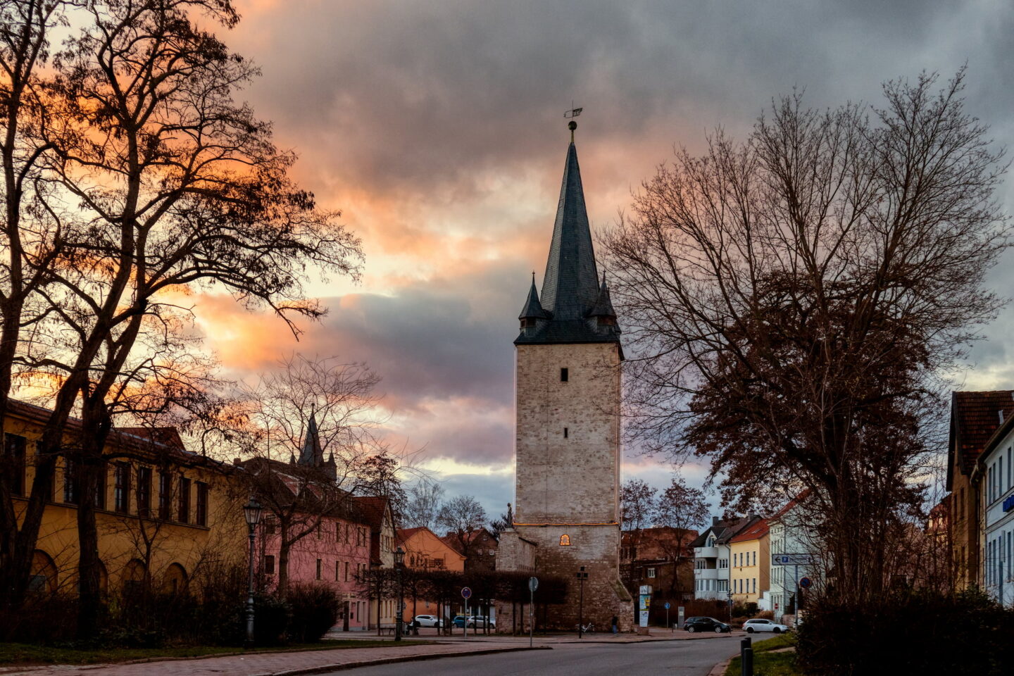 Ein hoher, schmaler Turm mit Spitzdach steht im Zentrum der historischen Stadt Aschersleben, umgeben von farbenfrohen Gebäuden. Kahle Bäume rahmen die Szene ein und am Himmel ist ein dramatischer Sonnenuntergang zu sehen, der die Gebäude und Wolken in einen warmen Glanz taucht. Eine Straße führt zum Turm.