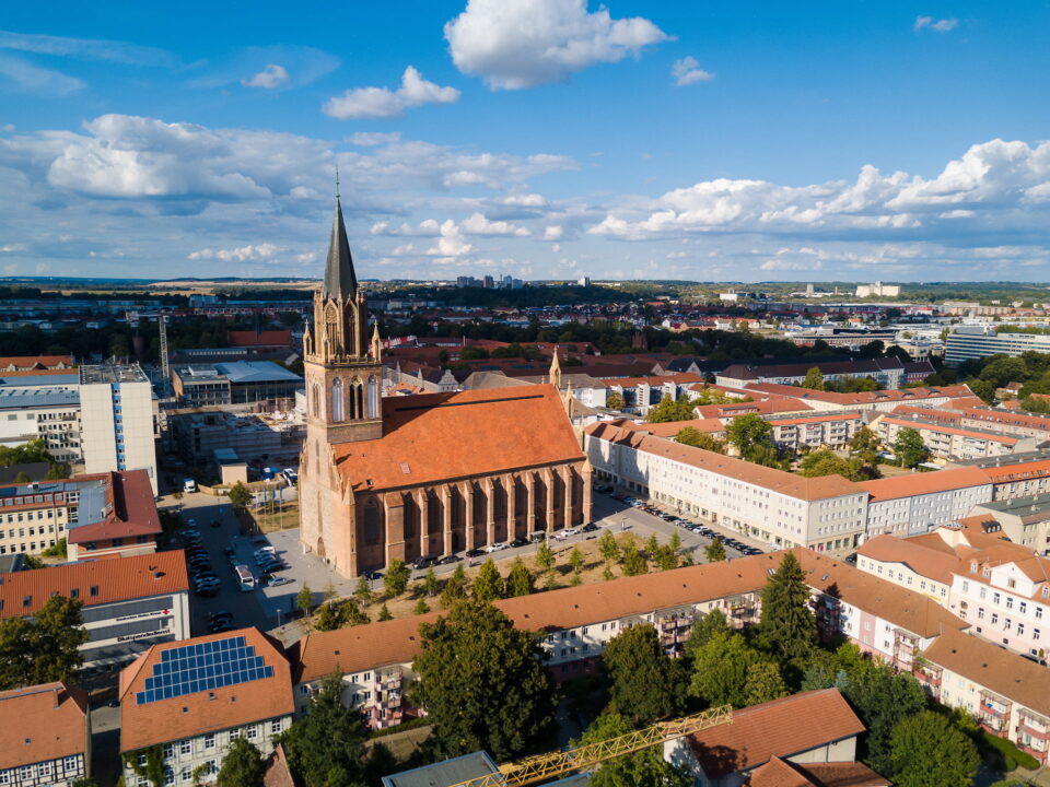 Luftaufnahme der malerischen Stadt Neubrandenburg mit einer markanten Kirche mit hohem Turm und roten Ziegeldächern. Zu den umliegenden Gebäuden gehören Wohn- und Geschäftshäuser, vor der Kulisse sanfter Hügel und eines teilweise bewölkten Himmels.