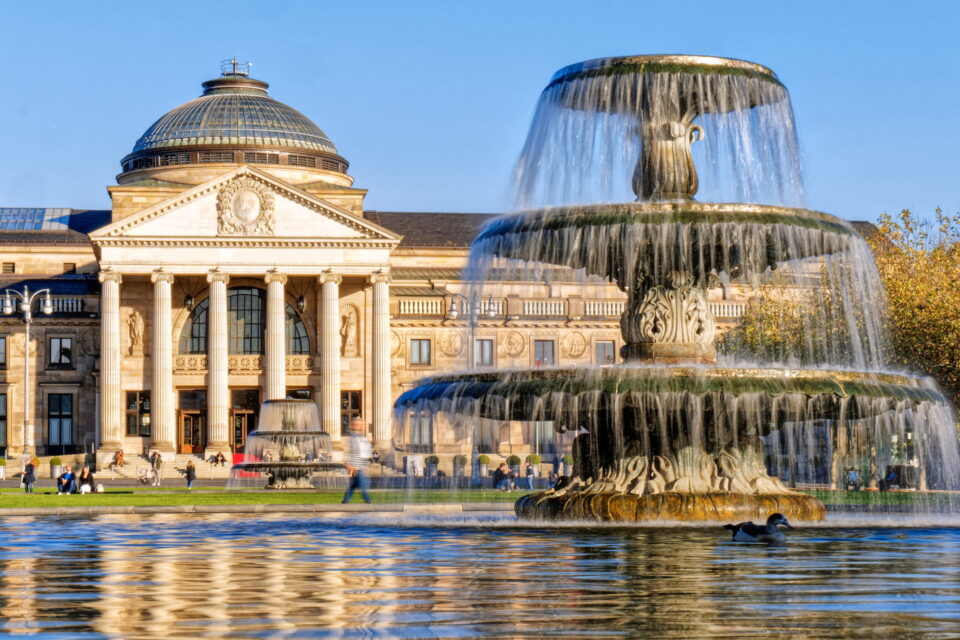 Im Vordergrund fließt ein eleganter, mehrstufiger Brunnen, im Hintergrund ein großes neoklassizistisches Gebäude mit Kuppel und Säulen. In der Nähe des Wiesbadener Wahrzeichens sieht man Menschen spazieren gehen und sich entspannen. Im Becken des Brunnens schwimmt eine Ente.