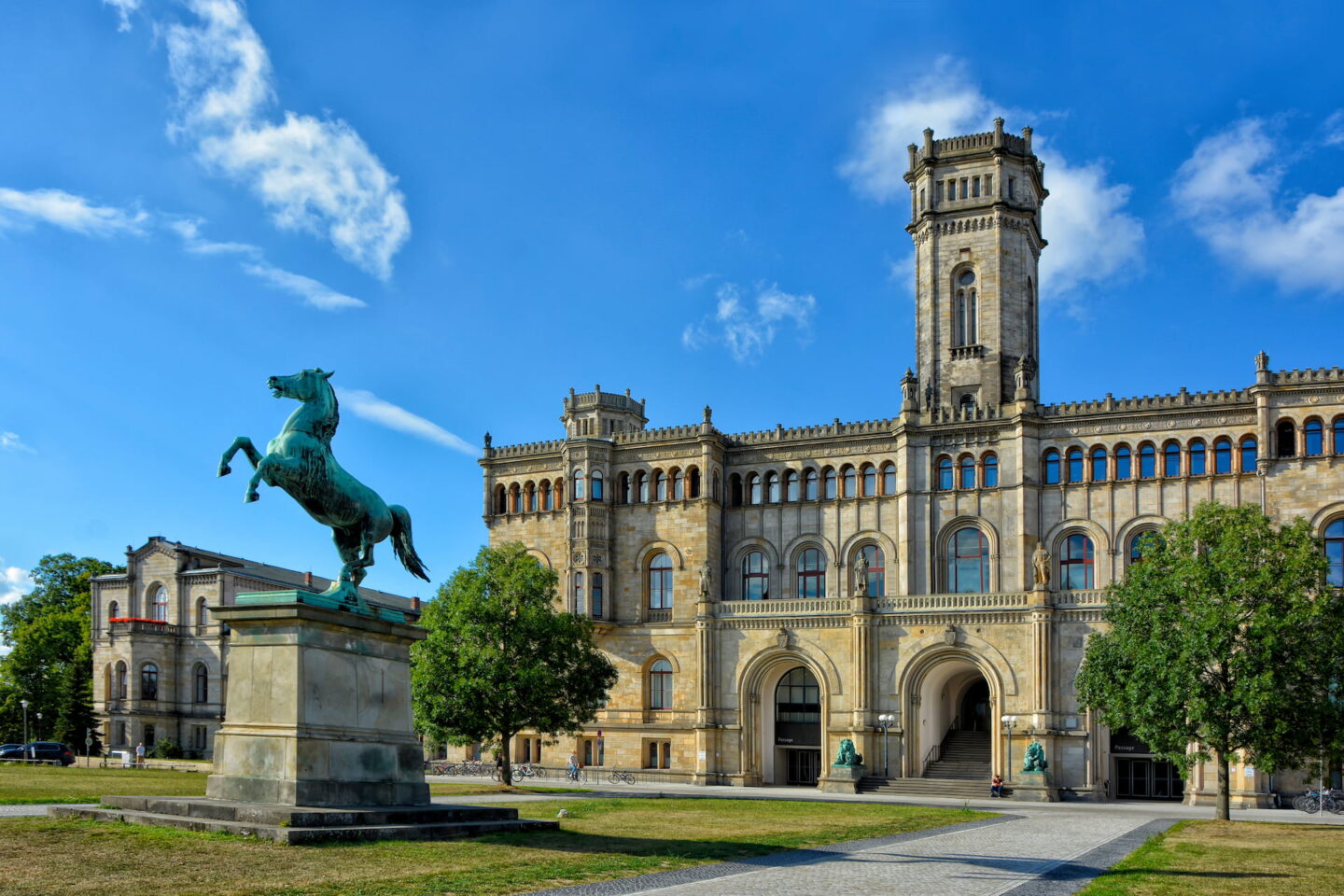 Ein historisches Gebäude in Hannover mit gewölbten Eingängen, zahlreichen Fenstern und einem zentralen Turm unter blauem Himmel. Vor dem Gebäude steht eine grüne Statue eines sich aufbäumenden Pferdes auf einem Sockel, im Vordergrund sind Bäume und ein Weg zu sehen.