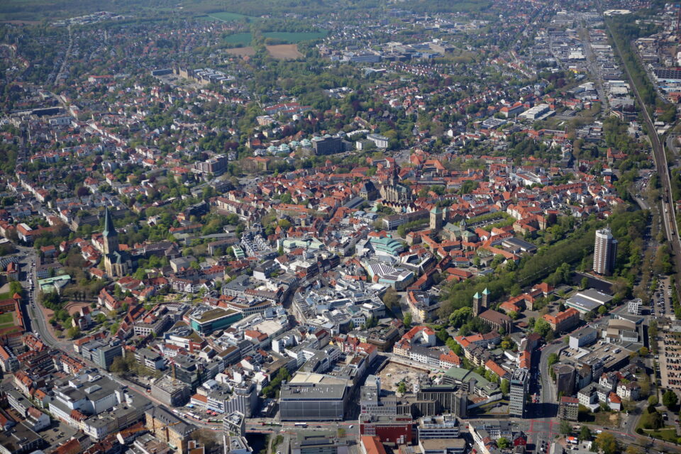 Luftaufnahme von Osnabrück mit einer Mischung aus historischer und moderner Architektur. Die Stadt hat dicht gedrängte Gebäude, von Bäumen gesäumte Straßen, Parks und offene Grünflächen. Die Landschaft umfasst eine markante Kathedrale sowie verschiedene Wohn- und Gewerbegebiete.