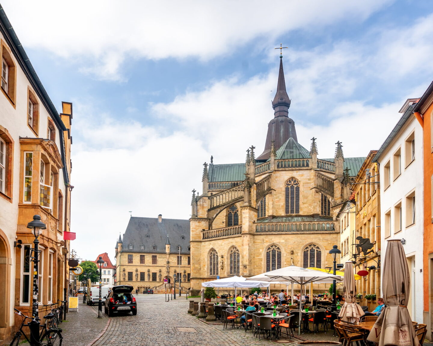 Eine Kopfsteinpflasterstraße in Luxemburg-Stadt mit Sitzgelegenheiten im Freien. Die neugotische Kathedrale Notre-Dame ragt prominent unter einem blauen Himmel hervor, umgeben von historischen Gebäuden, die an die in Osnabrück erinnern. Ein paar Menschen sind zu sehen und verleihen der malerischen Szenerie Leben.