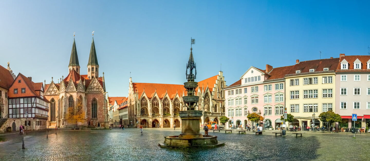 Panoramablick auf Braunschweigs historischen europäischen Stadtplatz mit einem zentralen Brunnen, alten Fachwerkhäusern, einer großen Kirche mit zwei Türmen und mehreren farbenfrohen mehrstöckigen Gebäuden unter einem klaren blauen Himmel. Fußgänger gehen spazieren und genießen den offenen Platz.