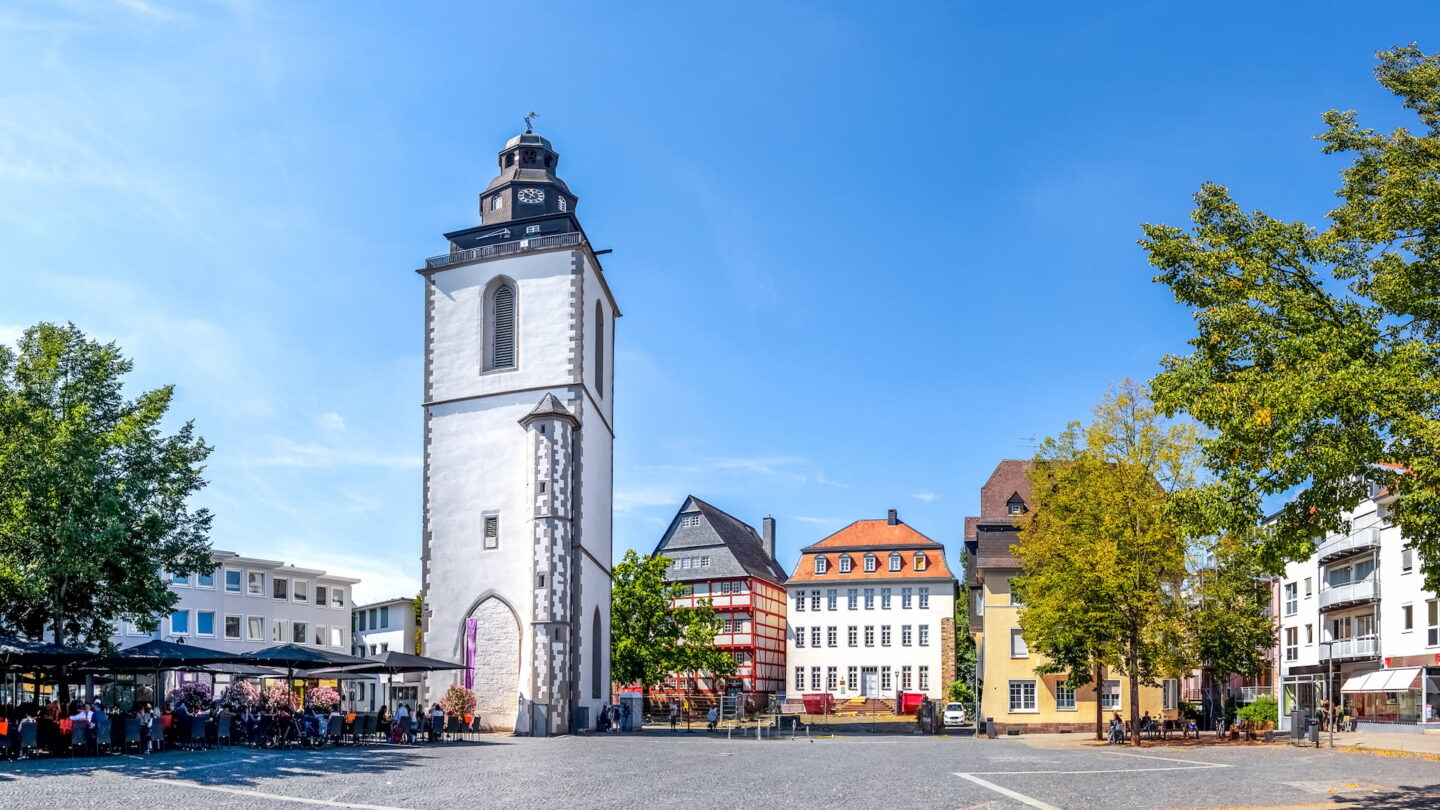 Ein sonniger Tag auf einem Stadtplatz mit dem ikonischen Eschenheimer Turm. Der weiße Turm mit seiner Uhr und dem Kirchturm ist von farbenfrohen historischen Gebäuden umgeben, die an die in Gießen erinnern. Bäume säumen den Platz, während sich Menschen unter einem klaren blauen Himmel in einem Straßencafé auf der linken Seite versammeln.