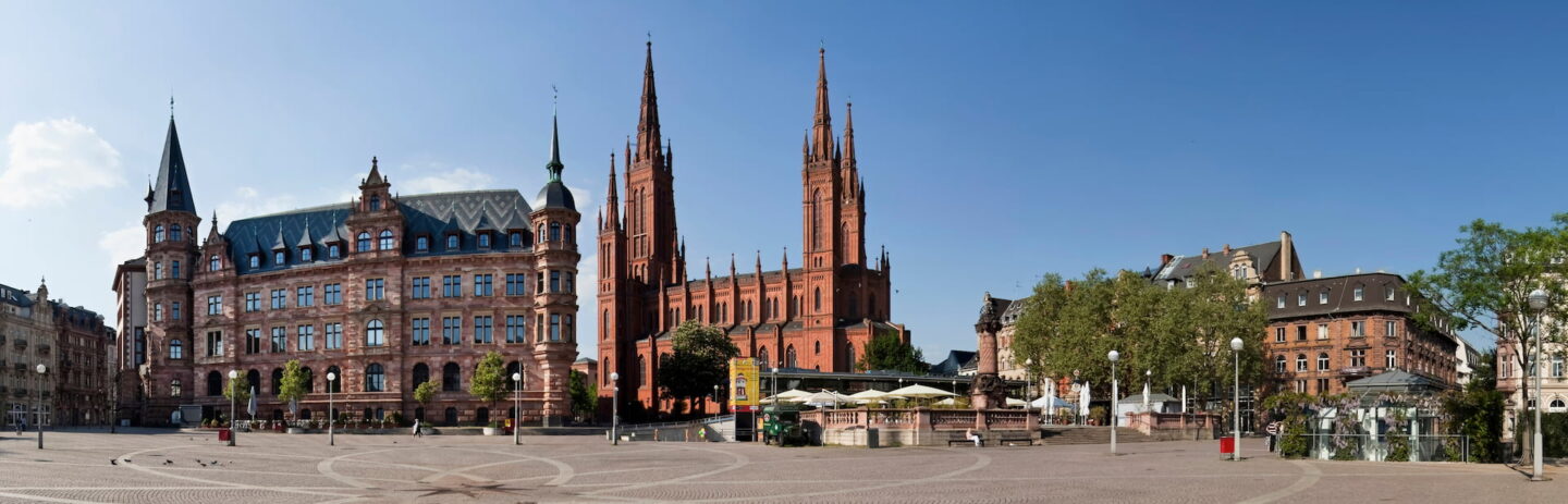 Ein großer, offener Platz mit gemustertem Steinpflaster in Wiesbaden ist von historischen Gebäuden umgeben. Auf der linken Seite ist ein großes Gebäude mit einem grünen Dach zu sehen, und in der Mitte steht eine große Kathedrale aus rotem Backstein mit zwei hohen Türmen unter einem klaren blauen Himmel. Rundherum sieht man Menschen und Cafés.